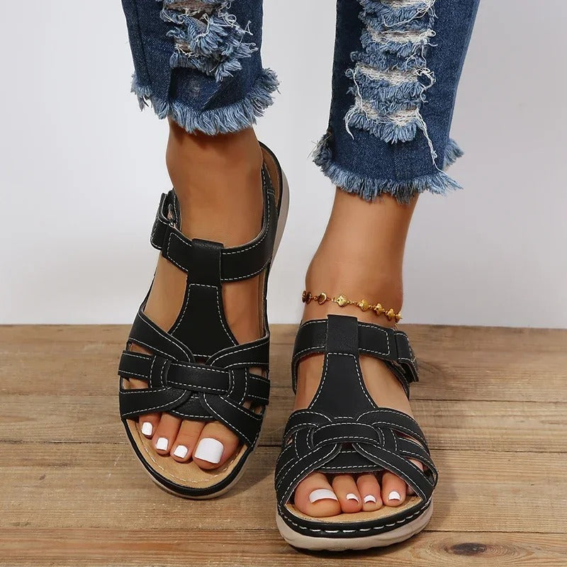 Black strappy sandals on woman's feet with white pedicure, paired with ripped blue jeans. Fashionable summer footwear on wooden floor.