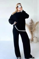 Woman in stylish black loungewear set with white trim, posing in minimalistic room with pampas grass and ceramic vases; trendy casual fashion outfit.