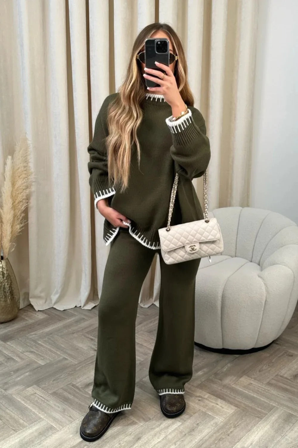 Woman in olive green knit sweater and pants set with white trim, holding a smartphone, standing next to a white quilted handbag on a chair.