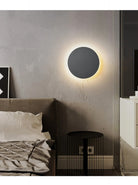 Modern bedroom with a round LED wall light, gray textured walls, minimalist bed, and black side table. Contemporary interior design, cozy ambiance.