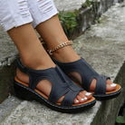 Women's navy blue open-toe sandals with white stitching, featuring a comfortable wedge heel and ankle strap, paired with ripped white jeans.