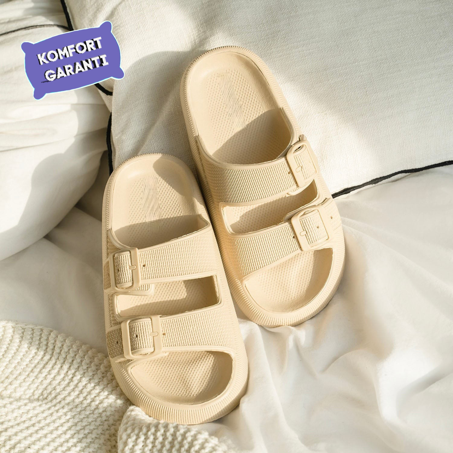 Beige slide sandals with dual adjustable straps on a white fabric background, featuring a "Komfort Garanti" label. Perfect for casual comfort footwear.