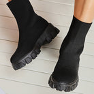 Black chunky platform sock boots with ribbed knit design, thick lug soles, and ankle-high fit on white wooden floor. Fashionable women's footwear.