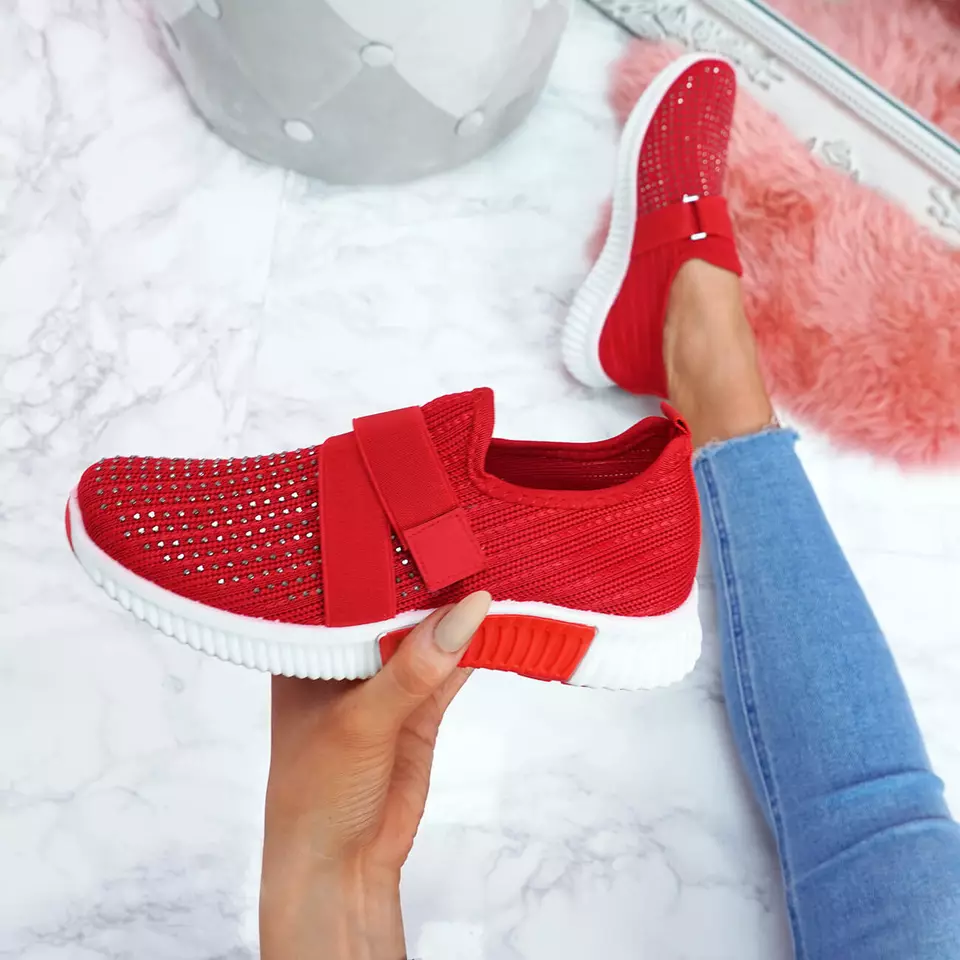 Red slip-on sneakers with white soles, featuring a breathable knit design and decorative rhinestones, held by a person wearing blue jeans on a marble floor.