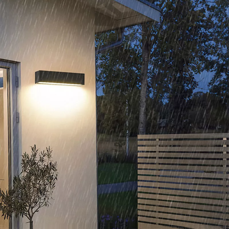 Modern outdoor wall light illuminating a house exterior during rain, featuring sleek black design. Ideal for garden, patio, or porch lighting solutions.