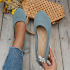 Women's blue knit ballet flats with pearl embellishment, held by a hand over a wooden floor, featuring a woven bag and yellow flowers.