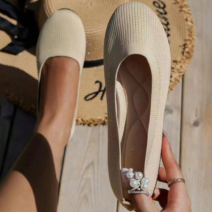 Beige knitted ballet flats with pearl embellishments, held in hand, on a wooden floor. Summer fashion footwear, stylish women's shoes.