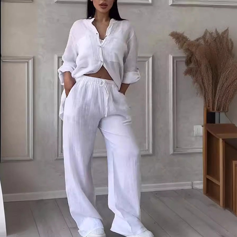 Woman wearing a stylish white linen outfit, featuring a button-up shirt and wide-leg pants, standing in a modern, minimalist room with pampas grass decor.