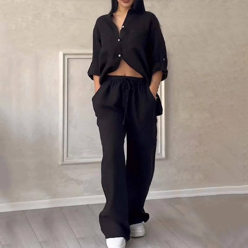 Woman wearing a stylish black two-piece lounge set with button-up top and wide-leg pants, standing indoors on light wood flooring.