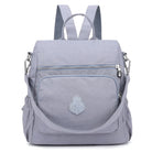 Light gray waterproof backpack with front zipper pocket, adjustable straps, and embroidered logo. Ideal for travel, school, or outdoor activities.