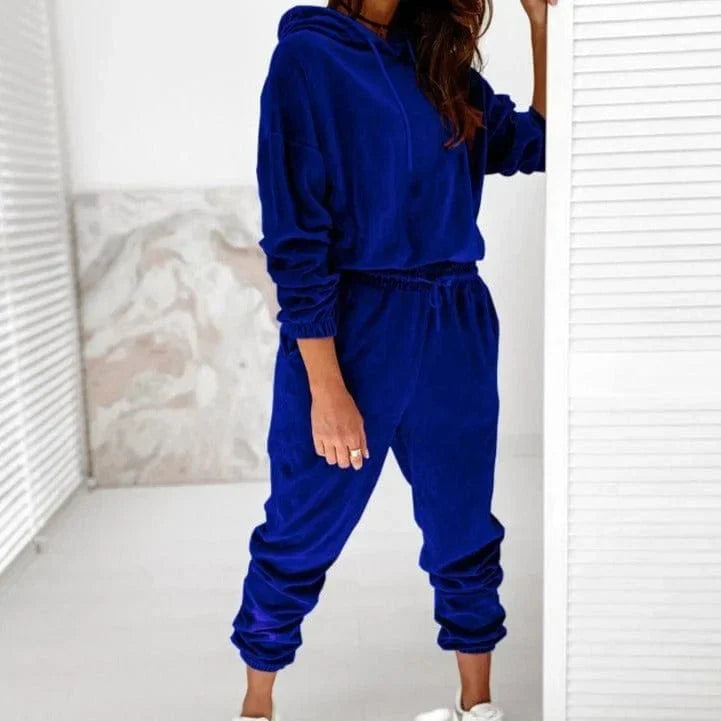 Woman wearing a blue velour sweatshirt and jogger set, standing in a modern, minimalist room. Fashionable loungewear outfit.