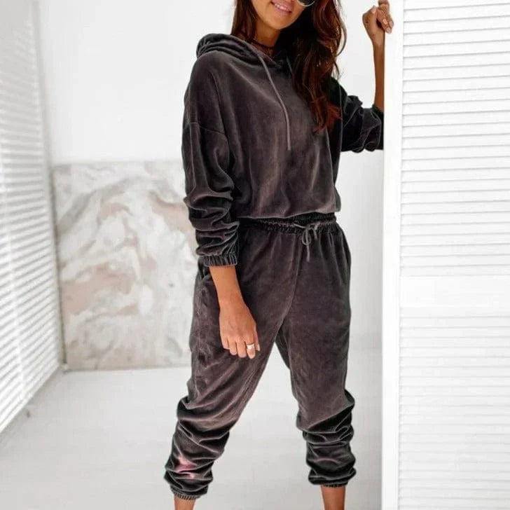 Woman wearing a stylish gray velour sweatshirt set with hoodie and jogger pants, standing indoors. Comfortable loungewear fashion outfit.
