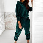 Woman wearing a green velour sweatshirt set, featuring a hoodie and jogger pants, standing indoors. Cozy loungewear fashion, comfortable style.