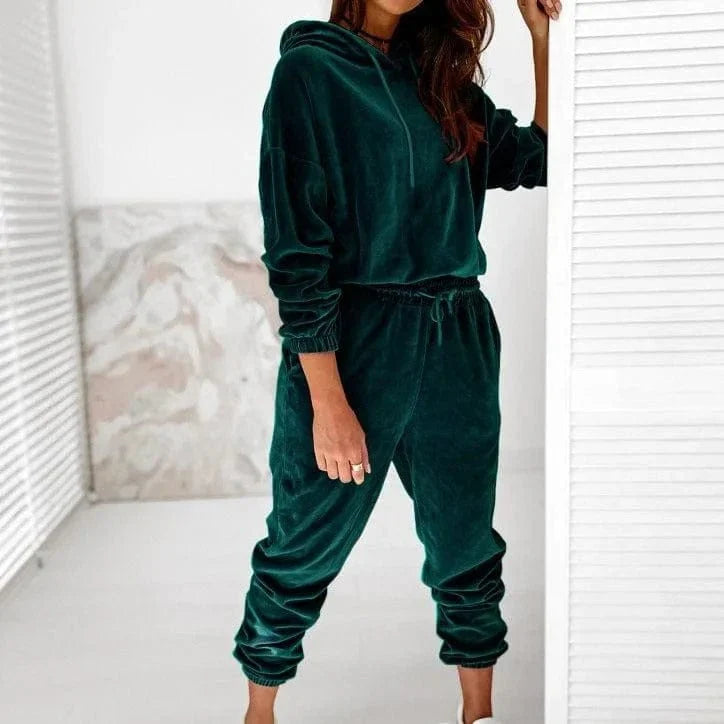 Woman wearing a green velour sweatshirt set, featuring a hoodie and jogger pants, standing indoors. Cozy loungewear fashion, comfortable style.