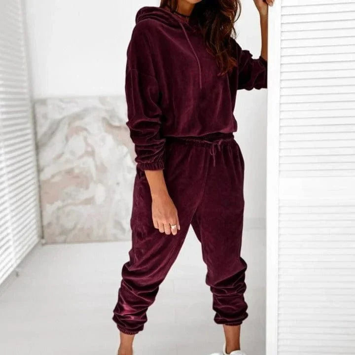 Woman wearing a burgundy velour tracksuit set, featuring a hoodie and joggers, standing in a modern, minimalist room. Fashionable loungewear outfit.