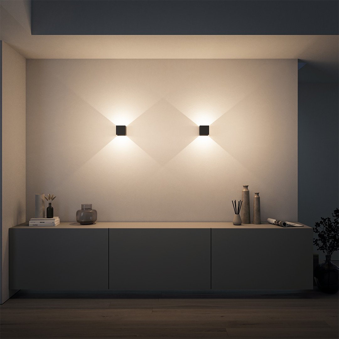 Modern minimalist interior with two geometric wall lights casting soft illumination above a sleek sideboard, featuring decorative vases and a plant.